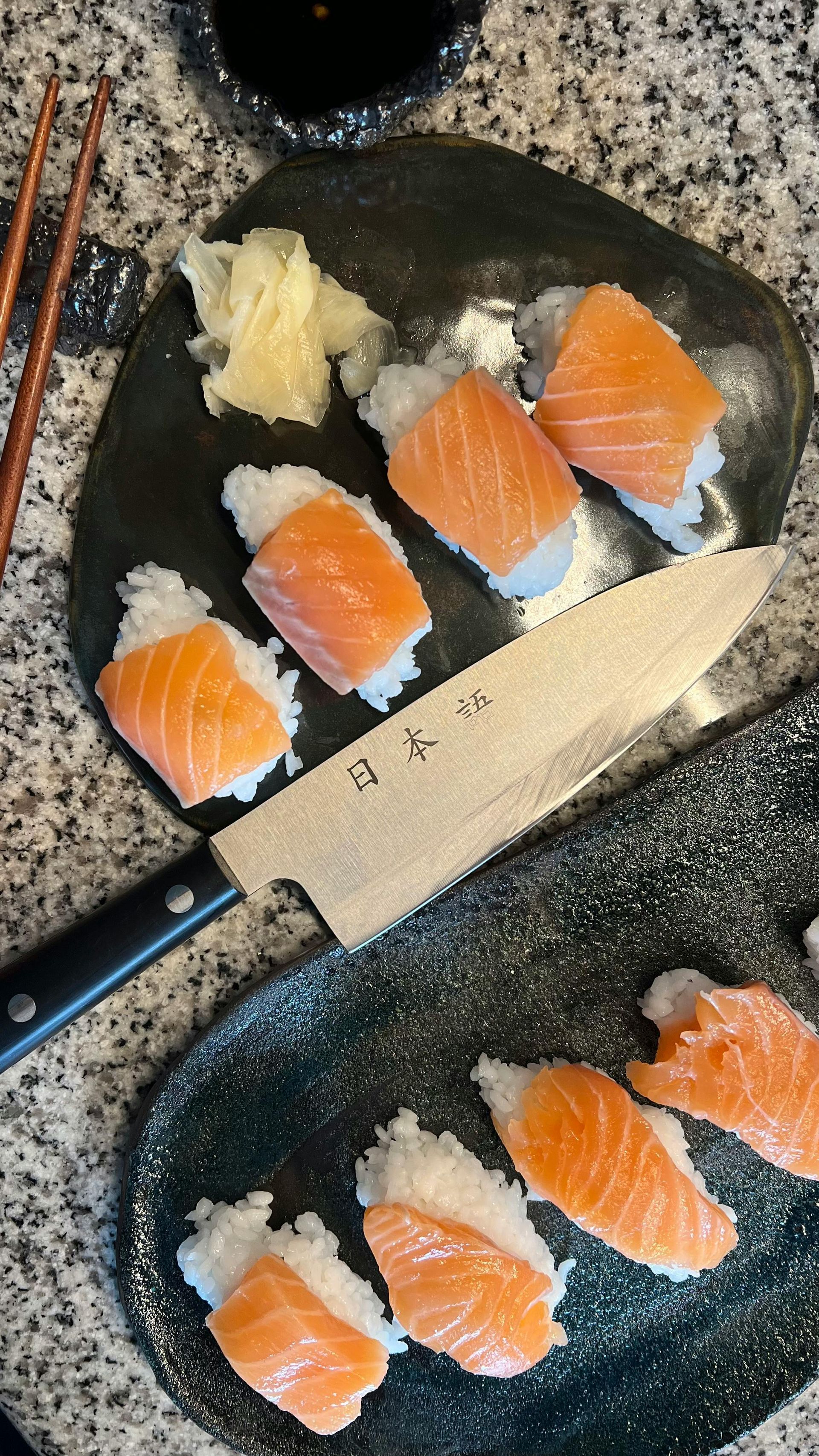Salmon sushi pieces on a dark plate with a knife, ginger, and chopsticks on a countertop.
