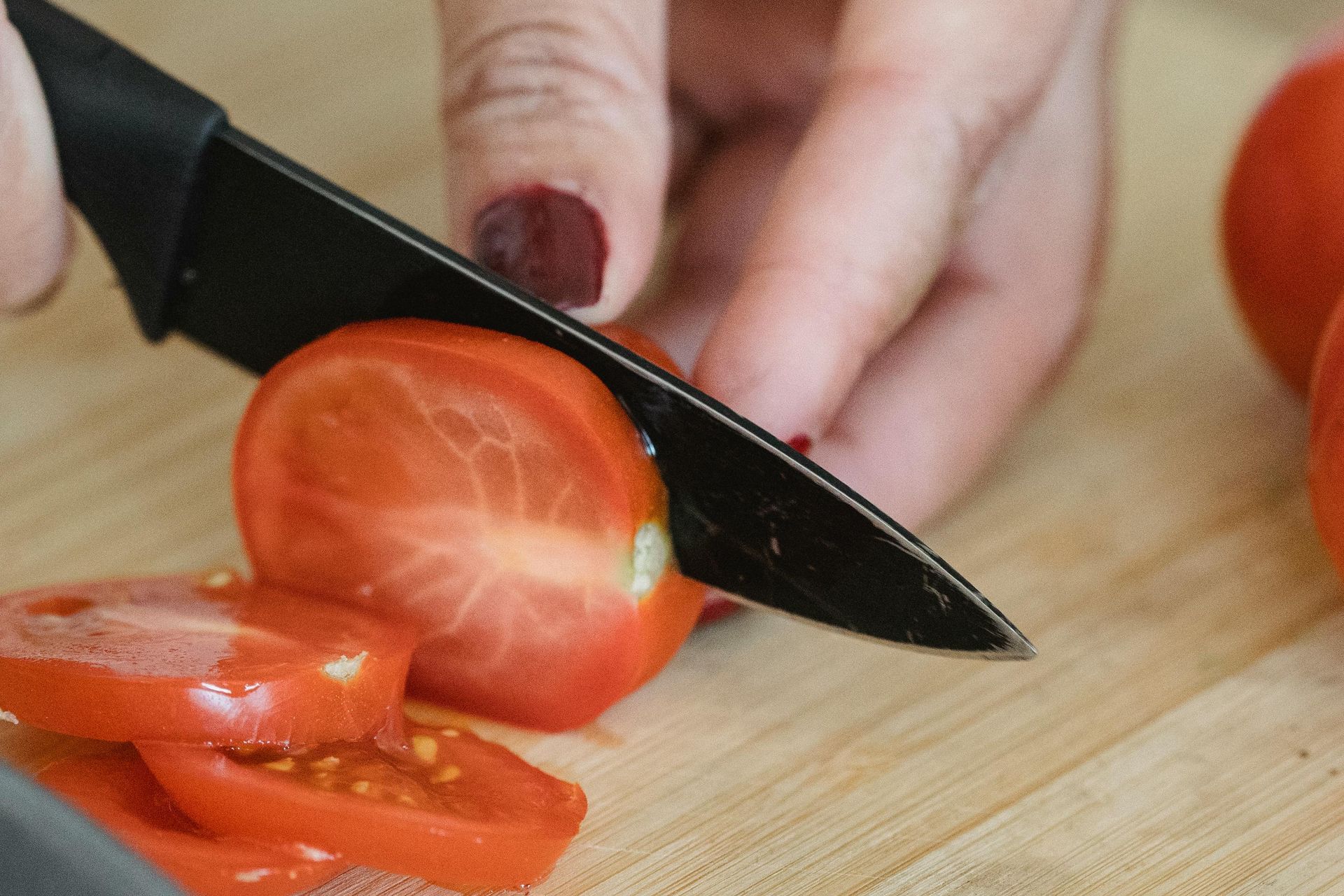 Hands slicing a red tomato on a cutting board with a black knife
