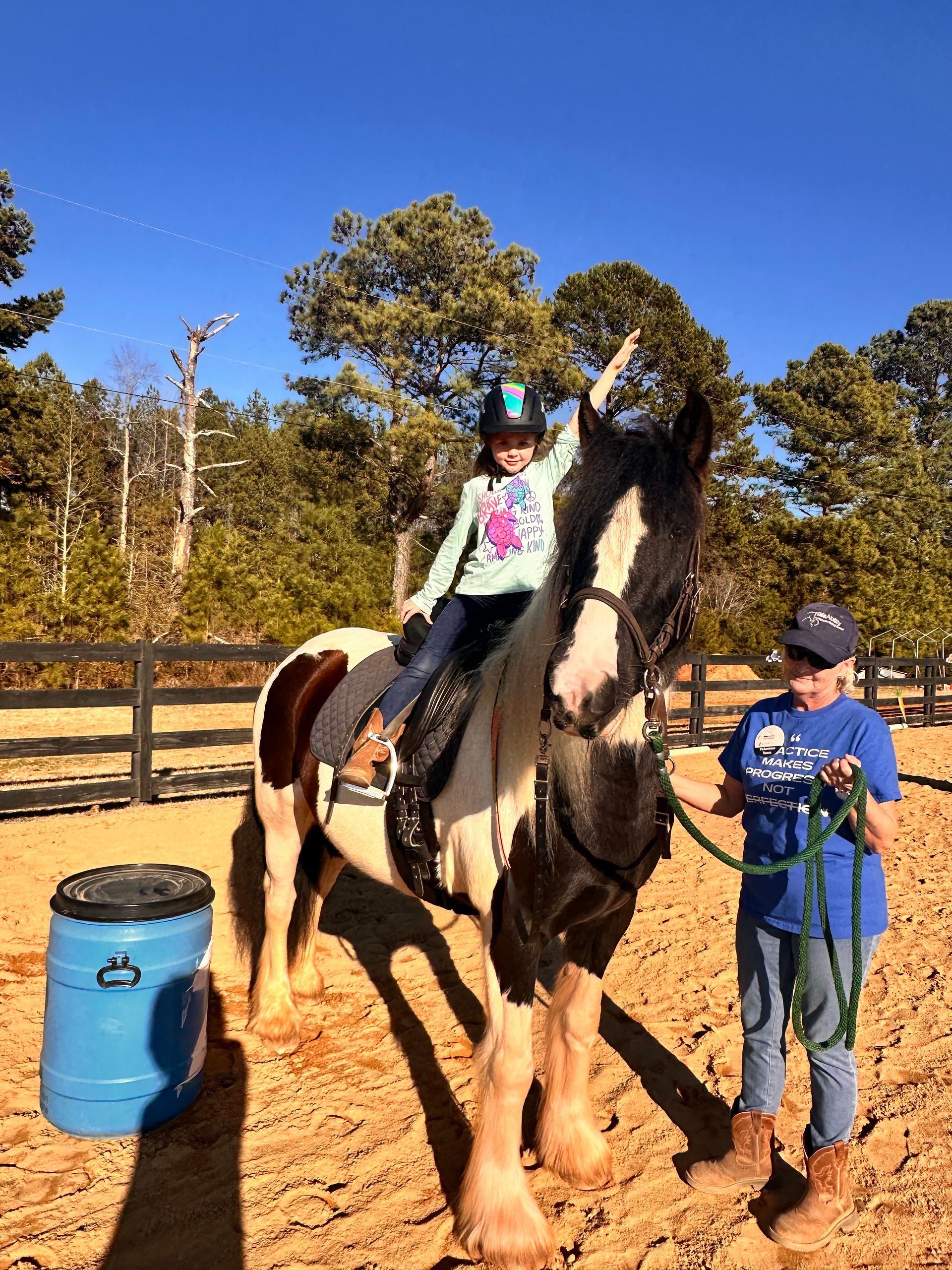 A young girl on a horse raises her arm. An adult stands next to them. Outdoor riding arena.