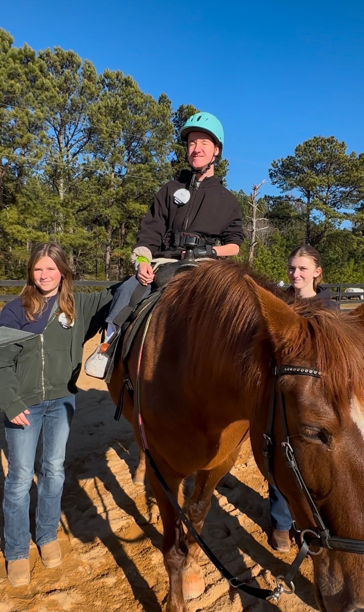 A person on a horse, flanked by two others, smiling. Outdoors, sunny, with trees in the background.