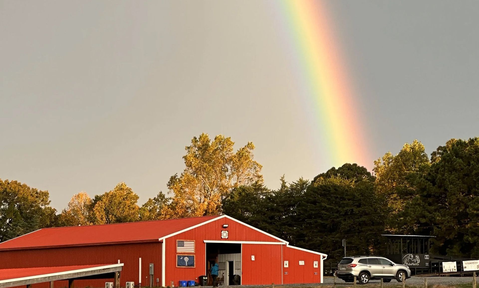 Red barn with a rainbow arcing across the sky. Trees and a car are in the background.