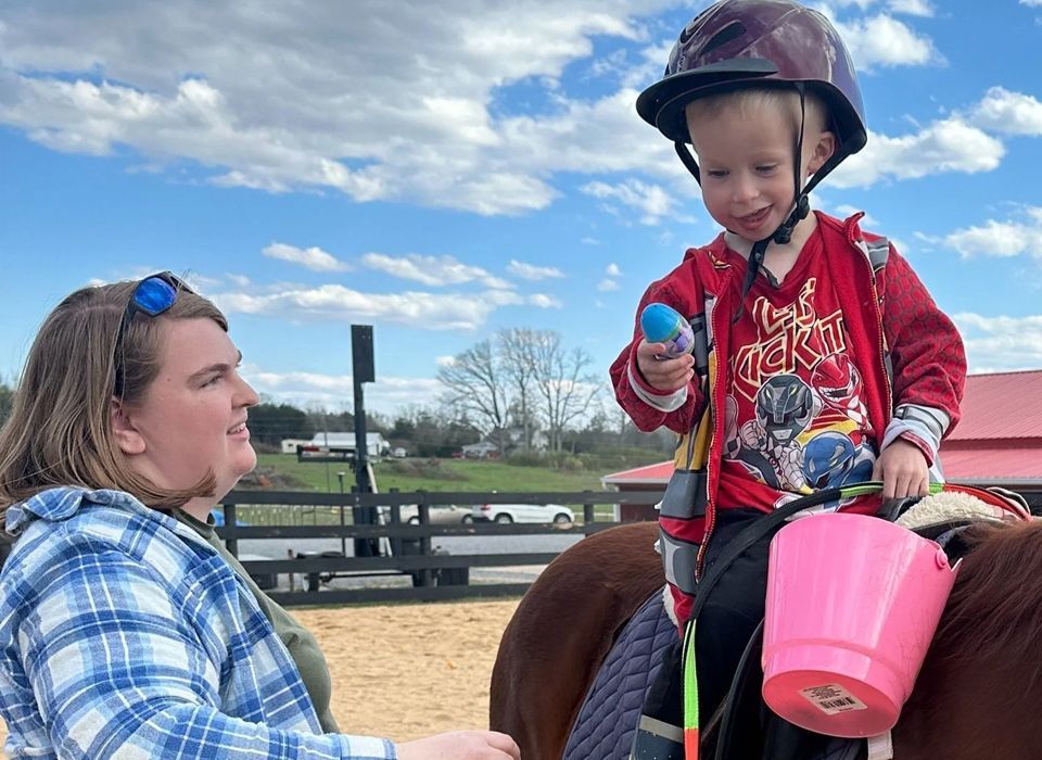 Girl riding a black and white horse with an instructor. Purple helmet, sunny day, barn in the background.