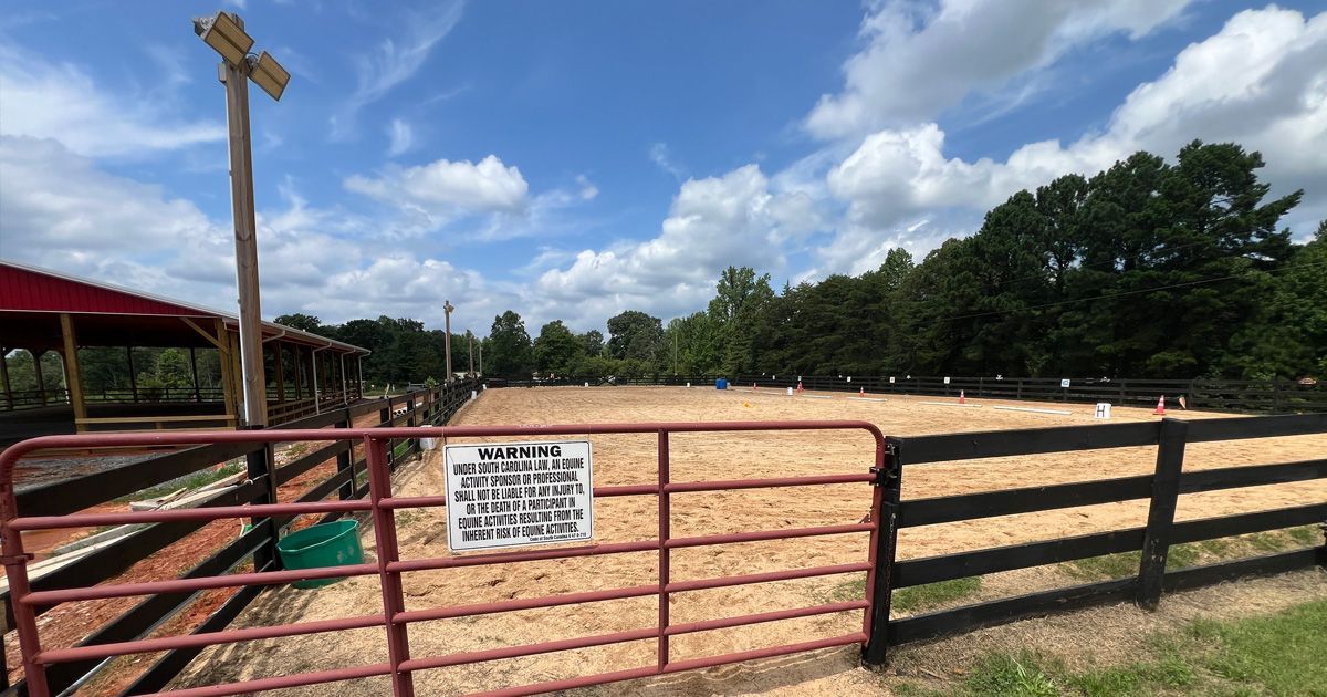Equestrian arena with tan sand, fenced in, under blue sky. Red gate, shelter, and sign in the foreground.