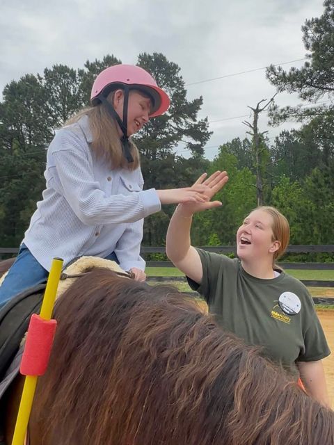 Woman on horse high-fives another woman, both smiling. Outdoors, trees in background.