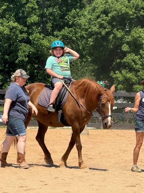 Boy with blue helmet on horse, being led by two adults in an outdoor riding arena; he smiles.