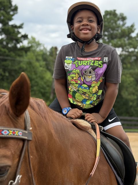 Boy in helmet, smiling, riding a brown horse in an outdoor arena.