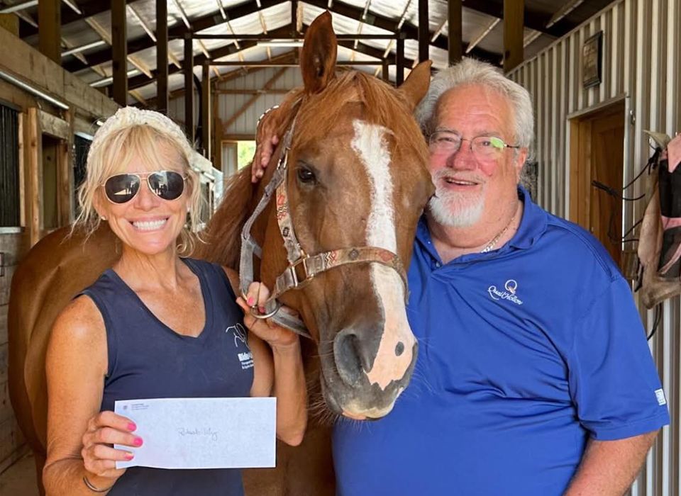 Woman, man, and a chestnut horse pose smiling. The woman holds a check. Inside a stable.