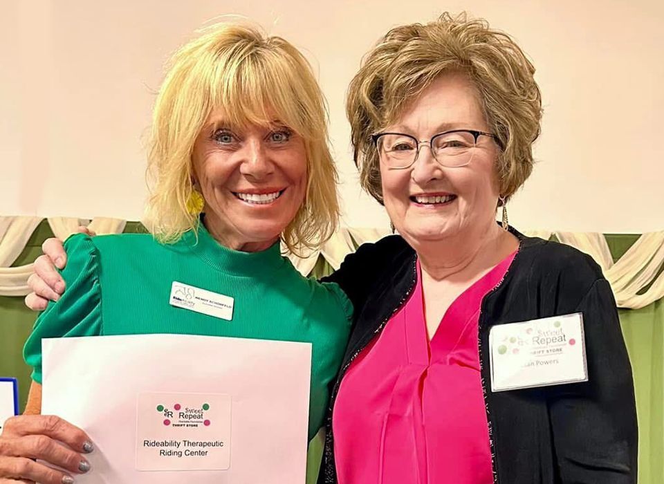 Two women smiling, one in green and holding a paper, the other in pink, indoors.