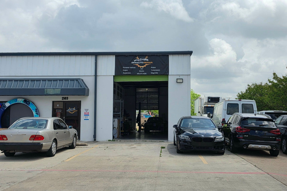 Auto repair shop exterior with parked cars and an open garage bay under cloudy skies | DFW Auto Repair