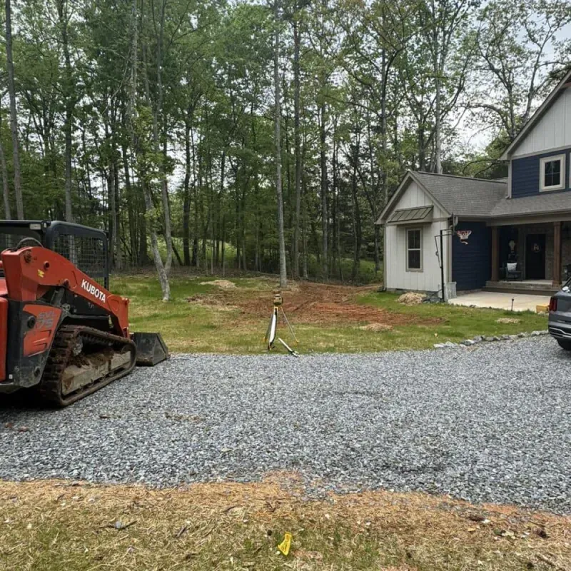 An orange skid steer loader sits on a gravel driveway next to a residential house and a wooded lot.