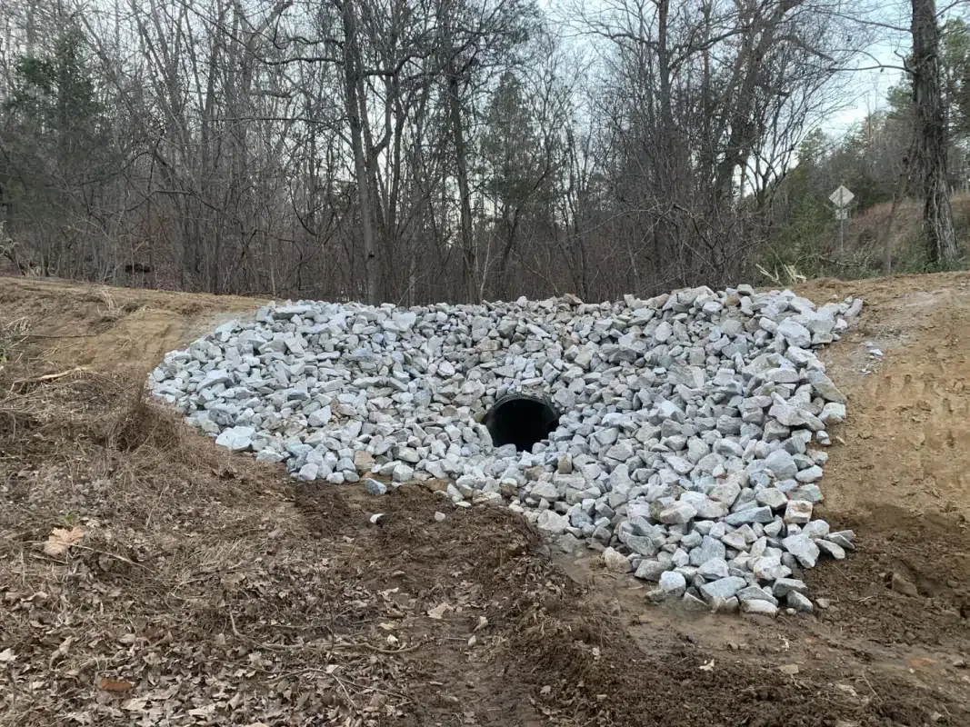 A drainage culvert surrounded by grey riprap stones at the base of a dirt embankment in a wooded area.