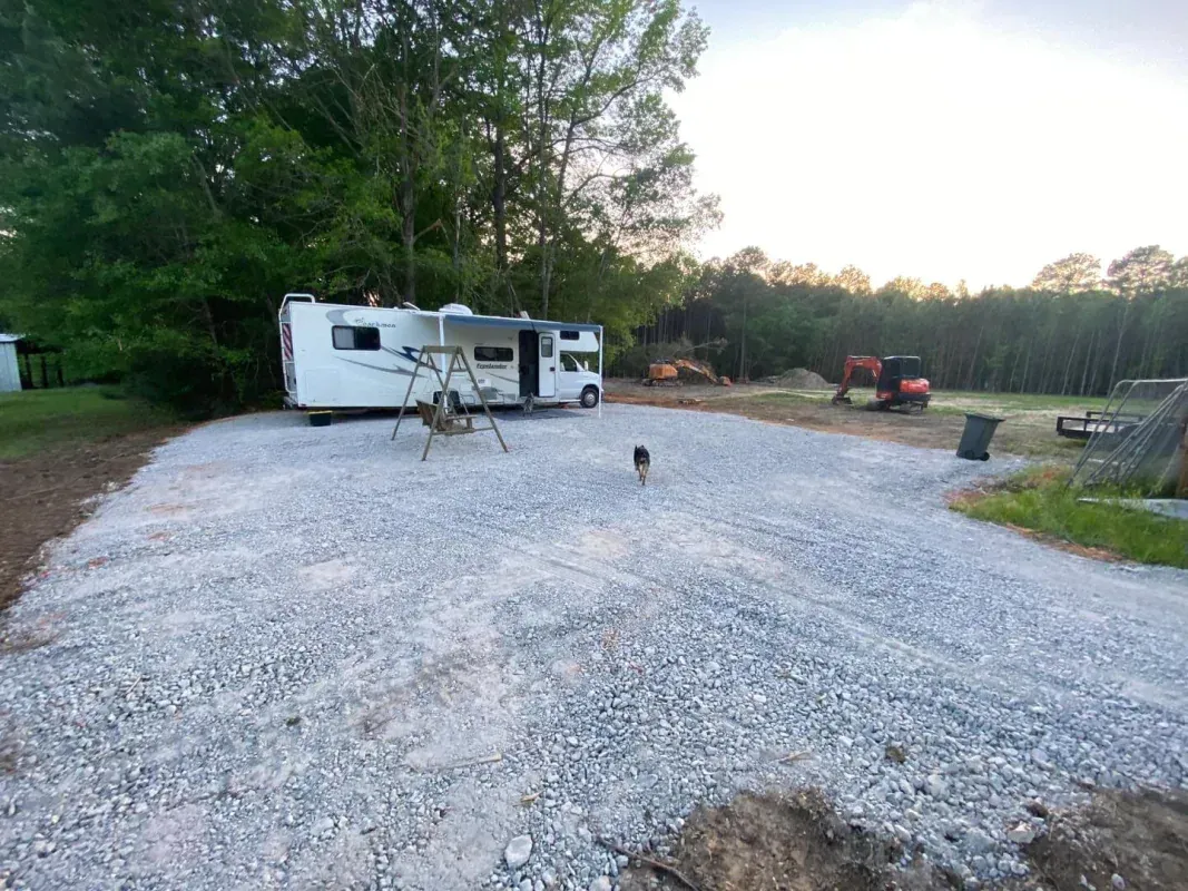 A white RV parked on a newly graveled lot next to trees, with a dog walking across the gravel and construction equipment.