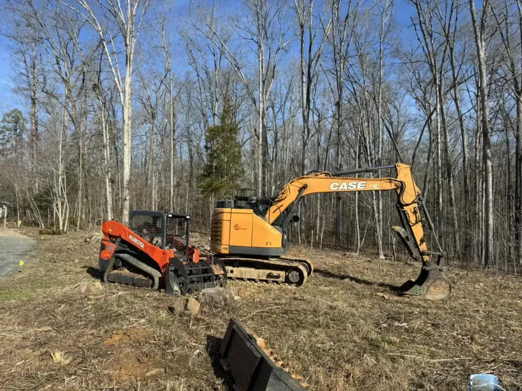 An orange skid steer and a yellow Case excavator sit on a cleared, grassy lot surrounded by bare trees.