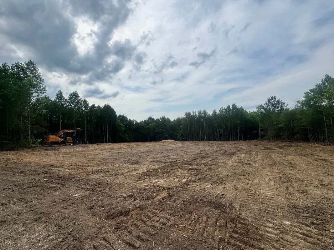 A clearing in a forest with brown, tilled earth under a cloudy sky, showing a construction vehicle on the left.