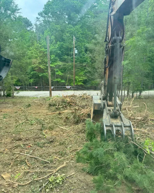 An excavator with a grapple attachment sits on a cleared, brush-covered lot near a road and a wooded area.