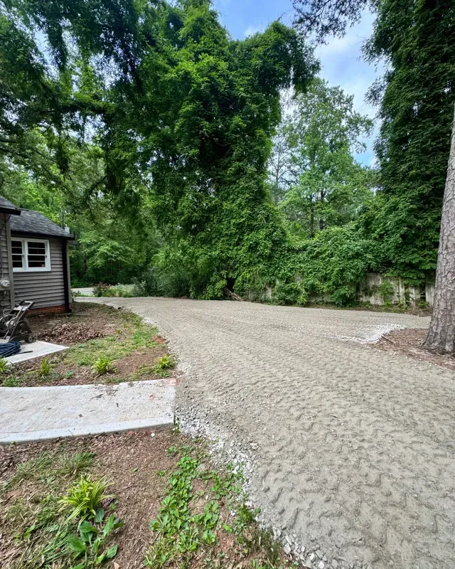 A freshly leveled gravel driveway curves toward a house next to dense green trees and foliage.