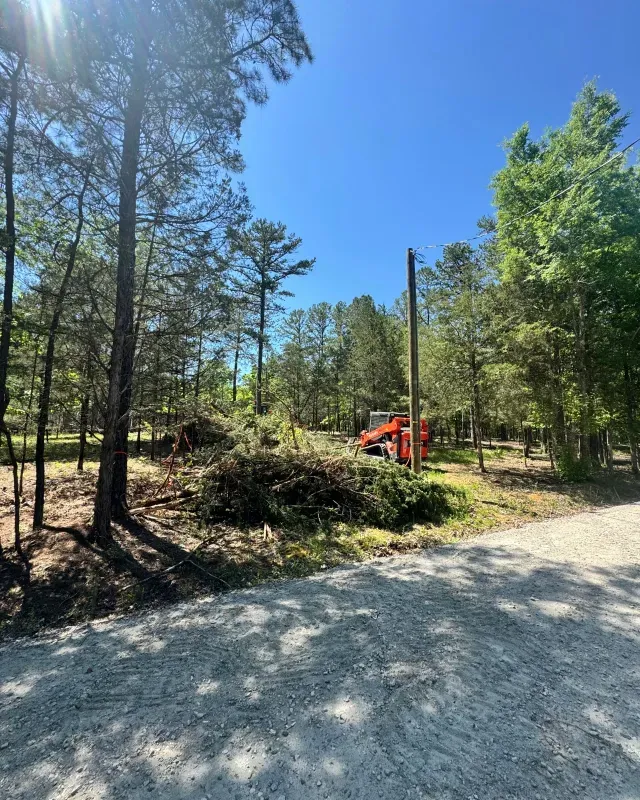 An orange piece of construction machinery sits on a dirt lot near trees and a gravel road under a bright blue sky.