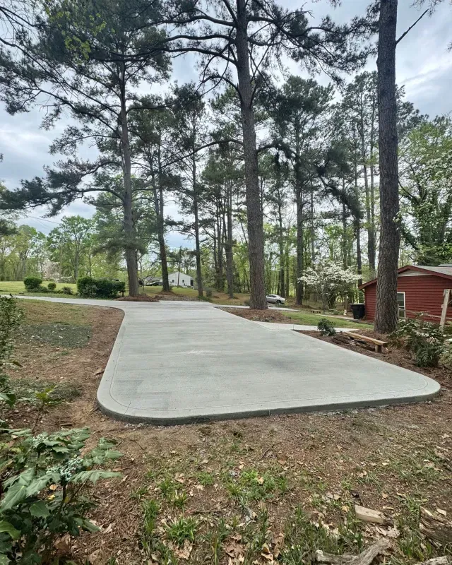 A freshly poured concrete driveway curves through a wooded yard toward a red house in the background under a cloudy sky.