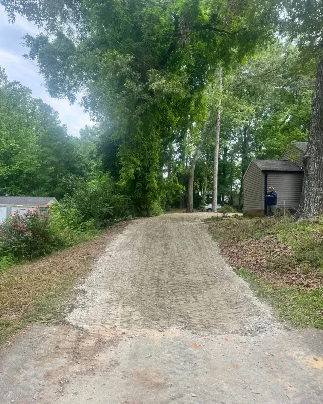 A gravel driveway slopes uphill between a house and trees, with a person visible near the side of the house.