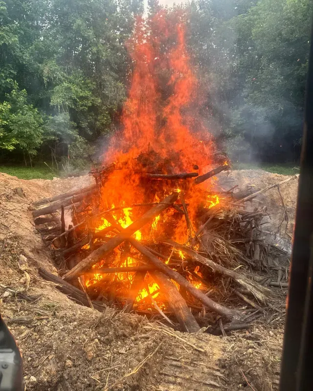 A large bonfire of logs and brush burning intensely within a dirt pit, framed by a forest background.
