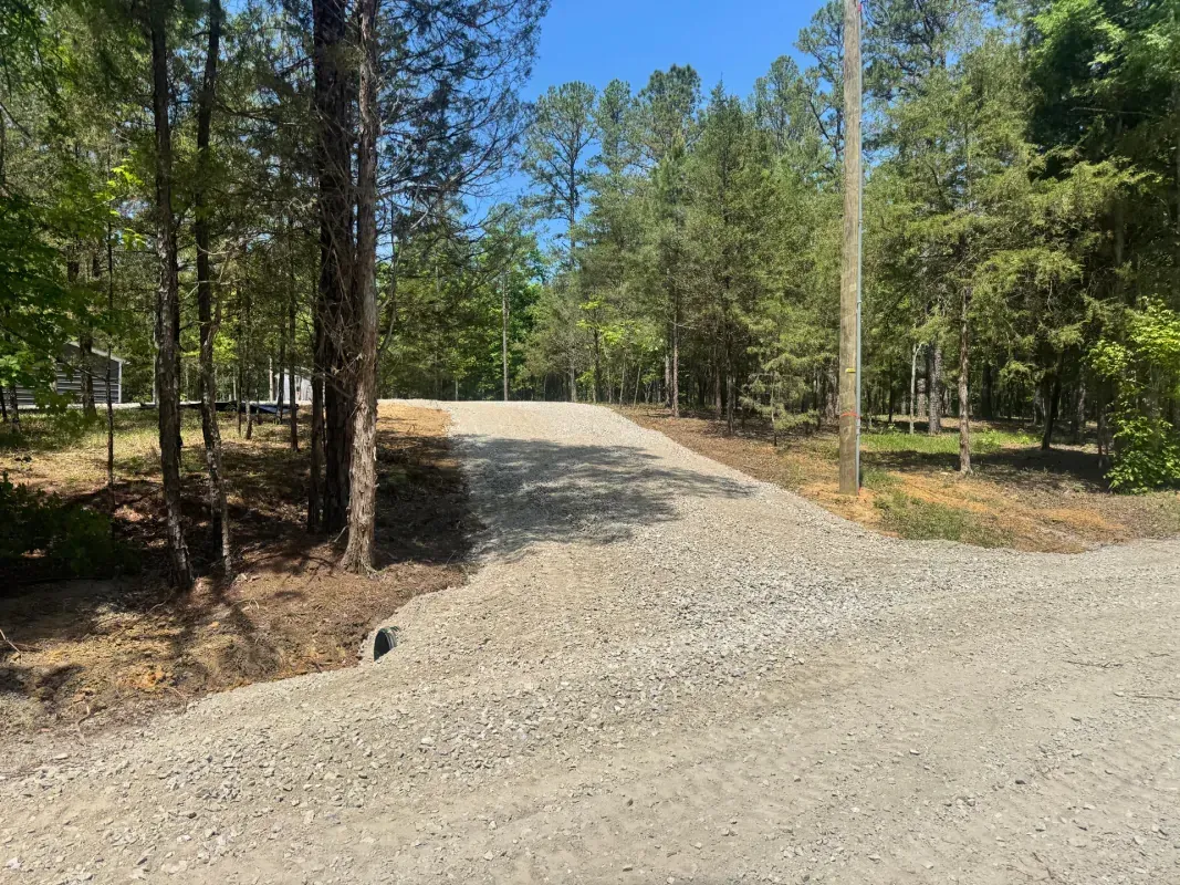 A gravel driveway extends from a dirt road into a wooded area on a sunny day, featuring a small drainage pipe.