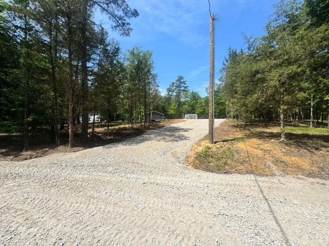 A gravel driveway leads through a wooded area toward a clearing under a bright blue sky.
