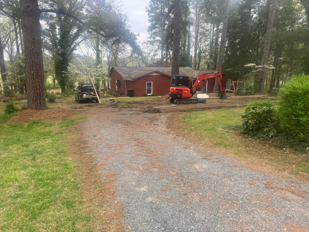 An orange excavator sits in front of a red house at the end of a gravel driveway surrounded by trees.