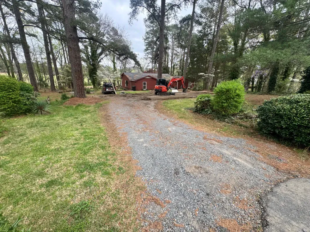A gravel driveway leads to a red house nestled among pine trees, with a small construction vehicle parked in front.