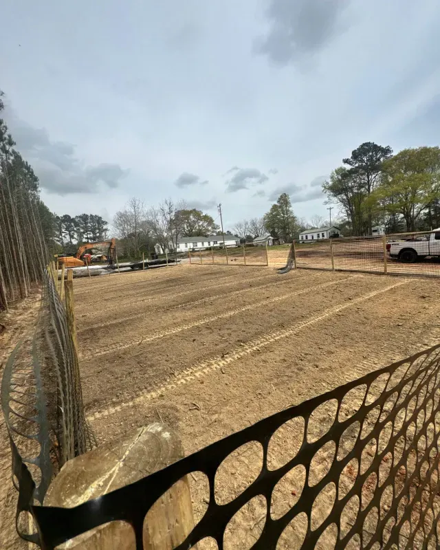 A fenced construction site with freshly tilled soil, tire tracks, heavy machinery in the distance, and trees under a cloudy sky.