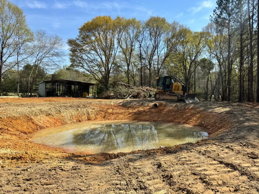 A bulldozer parks beside a newly excavated, muddy pond in a cleared, rural area with trees in the background.