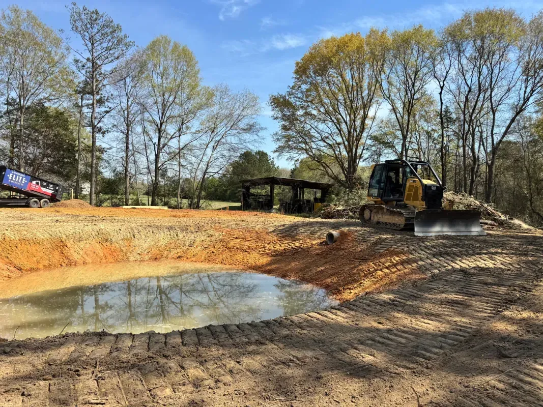 A yellow bulldozer sits beside a freshly excavated pond with orange soil in a wooded clearing under a clear blue sky.