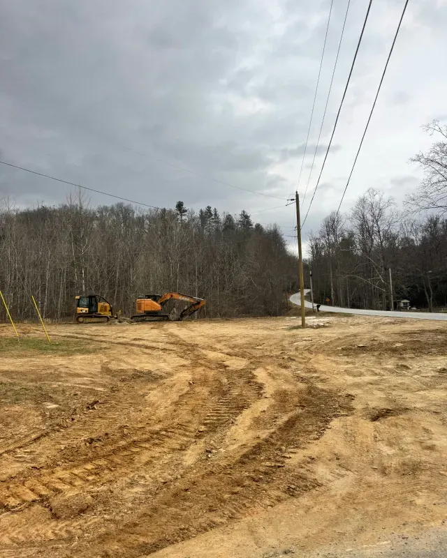 Two construction excavators parked on a cleared, dirt-covered lot near a road and a forest under a cloudy sky.