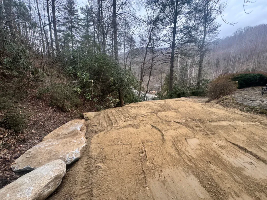 A dirt road descends through a wooded area lined with large boulders on the left, under an overcast sky.