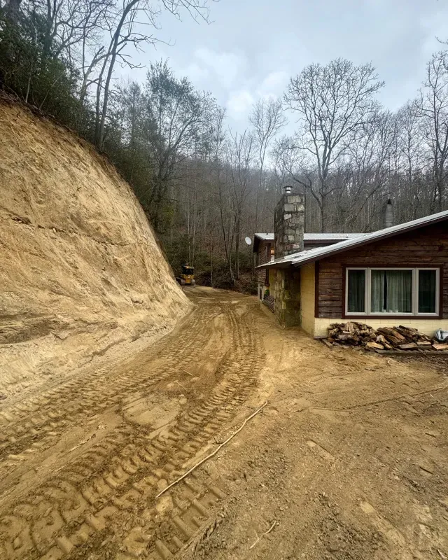 A dirt road with tire tracks curves past a hillside beside a wood-sided house with a stone chimney on a cloudy day.