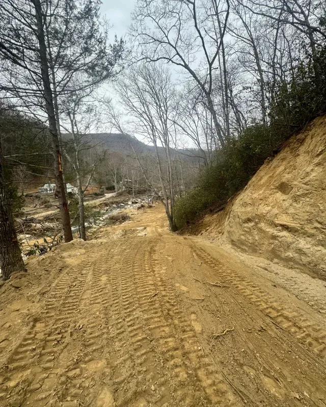 A dirt road with deep tire tracks slopes downward, flanked by a steep, raw earth embankment and bare trees.