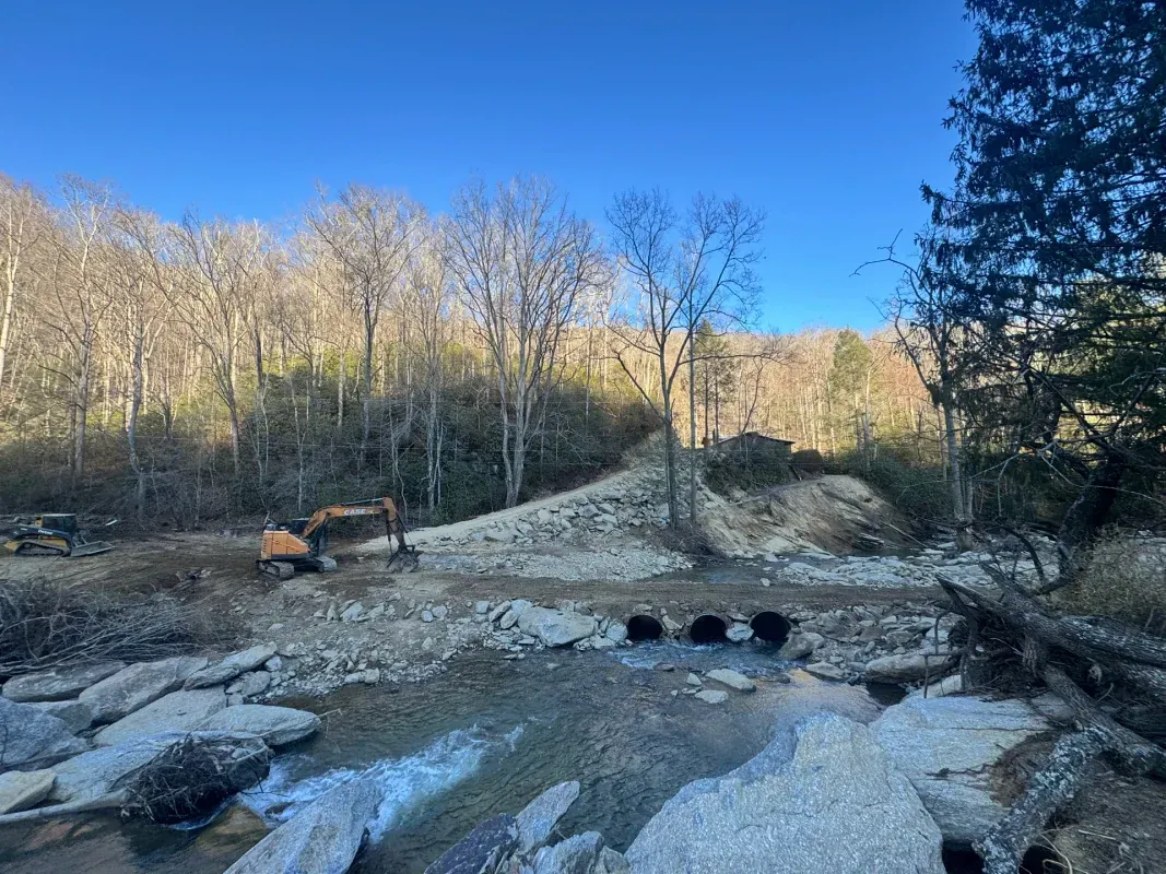 An excavator works on a small dirt road with culverts over a rocky stream in a wooded landscape under a clear blue sky.