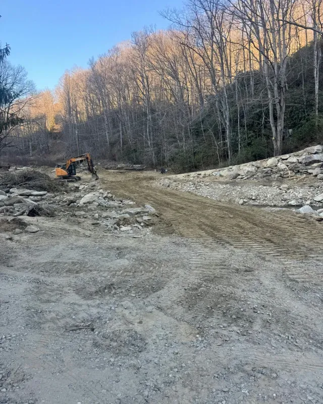 An excavator works on a dirt path in a wooded area with large rocks piled along the side.