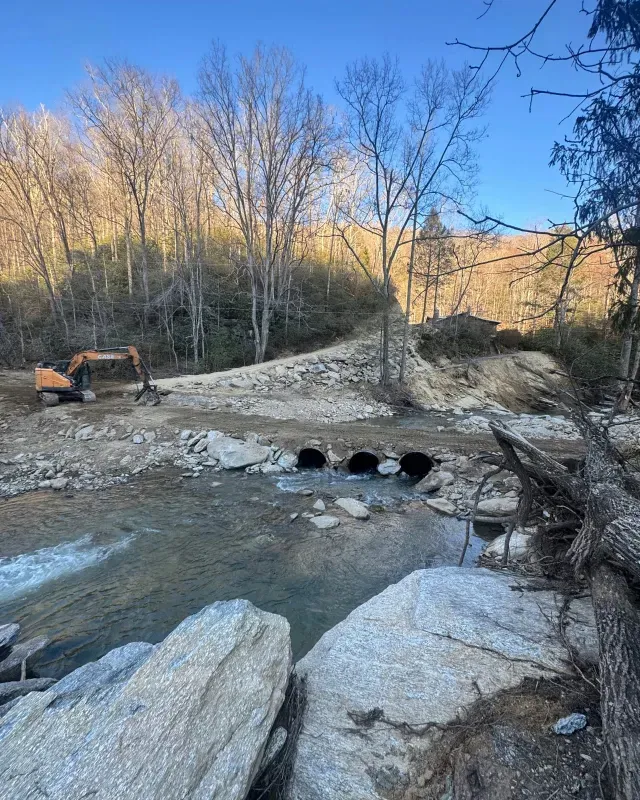 An excavator works on a rocky crossing over a stream with three culverts, set against a backdrop of bare autumn trees.