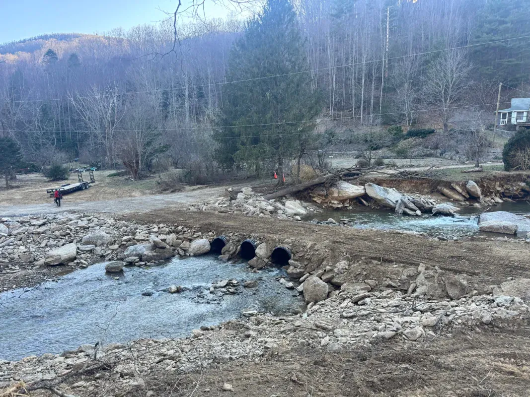 A dirt crossing with three culverts over a rocky stream, with eroded banks, forested hills, and workers in the distance.