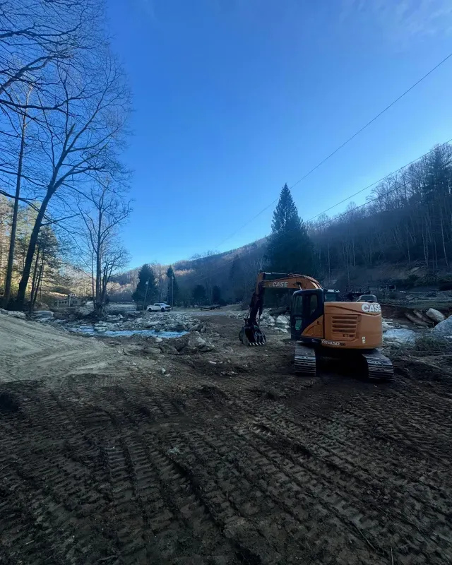 An orange excavator sits on a dirt road by a river in a hilly, wooded area under a clear blue sky.