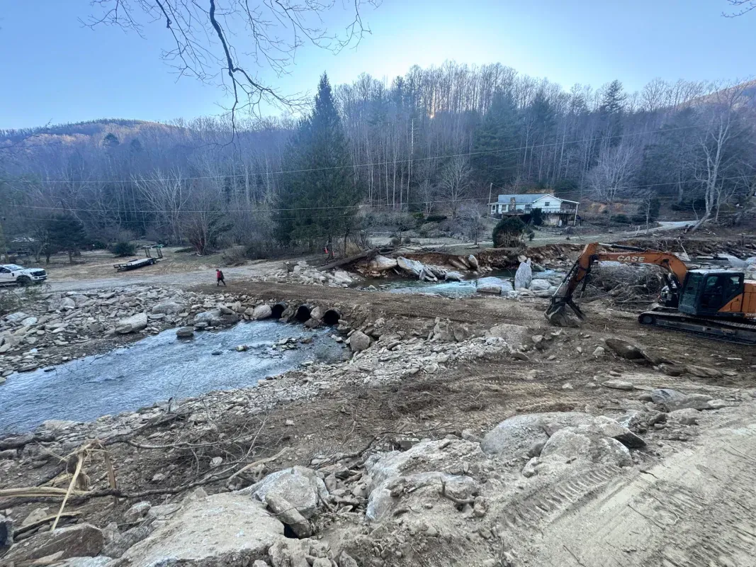 An excavator works on a rocky riverbank, moving earth to clear a path near large drainage pipes and a forested hillside.