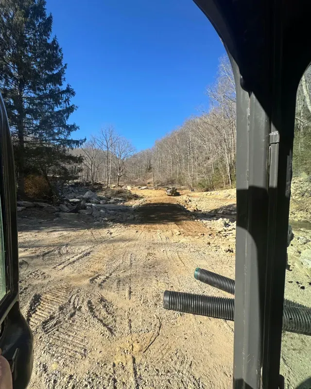 View from inside a cab showing a dirt construction path leading toward wooded hills under a clear blue sky.