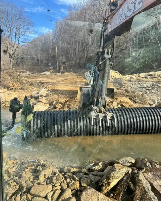 An excavator uses a thumb attachment to position a large, black corrugated plastic drainage pipe in a muddy stream.