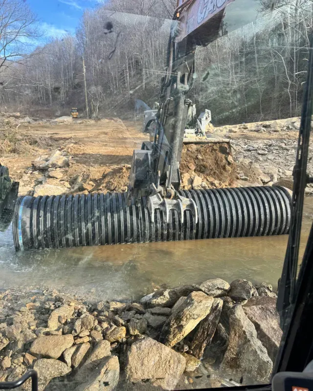 An excavator holds a large, black, corrugated plastic pipe over a shallow stream at a construction site.