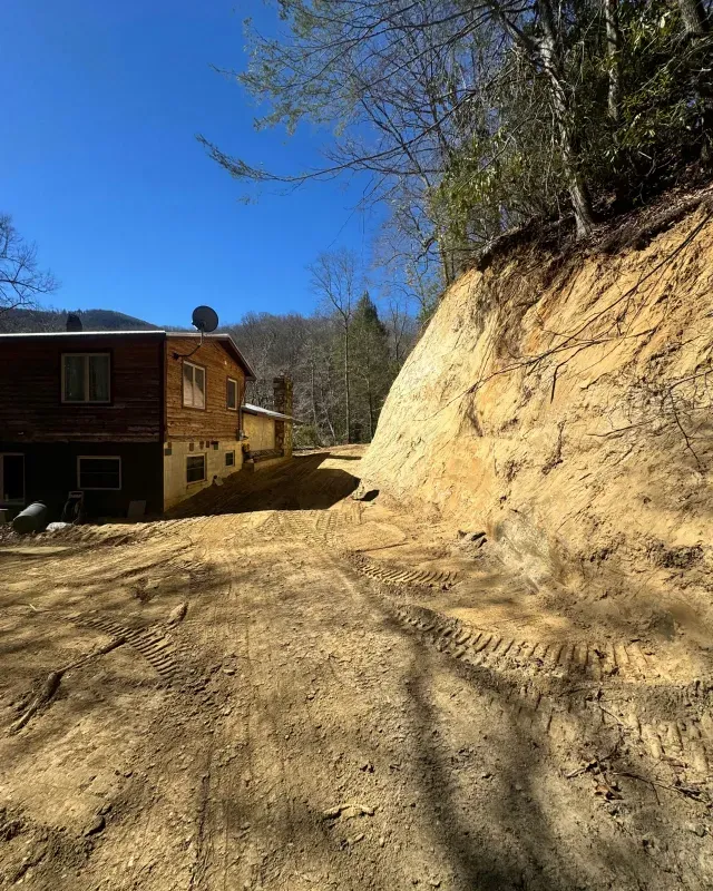 A two-story house stands beside a newly excavated, steep dirt embankment under a clear blue sky.
