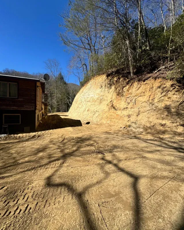 A dirt path winds beside a cabin toward a steep, freshly excavated hillside beneath a clear blue sky.