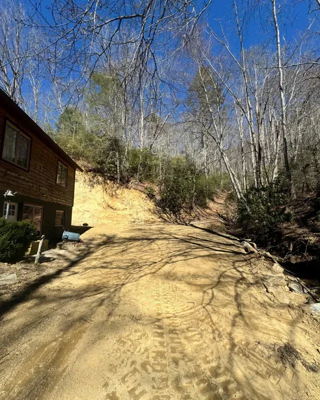 A dirt driveway winds upward past a wooden house toward a wooded hillside under a bright blue sky.