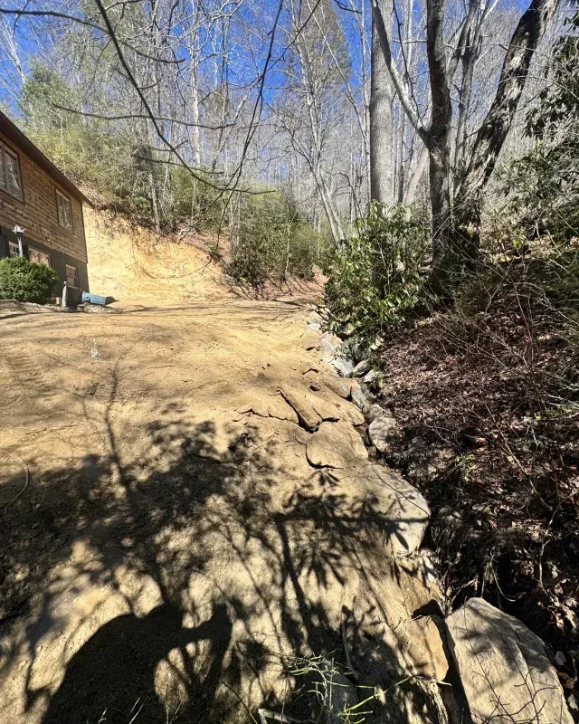 A dirt driveway beside a hillside, with a wooden house on the left, trees in the background, and a rock-lined slope.