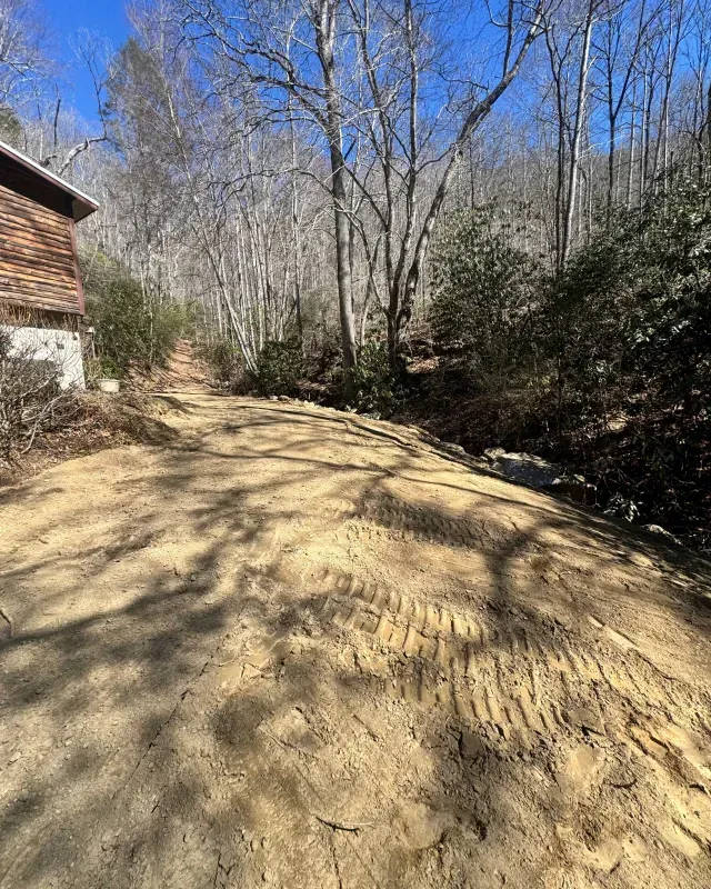 A gravel driveway leads past a house toward trees and a wooded hillside under a bright blue sky.