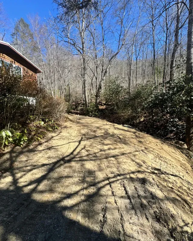 A sloping, freshly graded dirt driveway leads upward toward a wooden house nestled in a forest of bare trees.